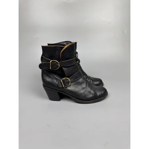 May include: A pair of black leather ankle boots with a stacked heel. The boots feature two adjustable straps with gold-colored buckles and a suede collar. The boots are worn and have a vintage look.