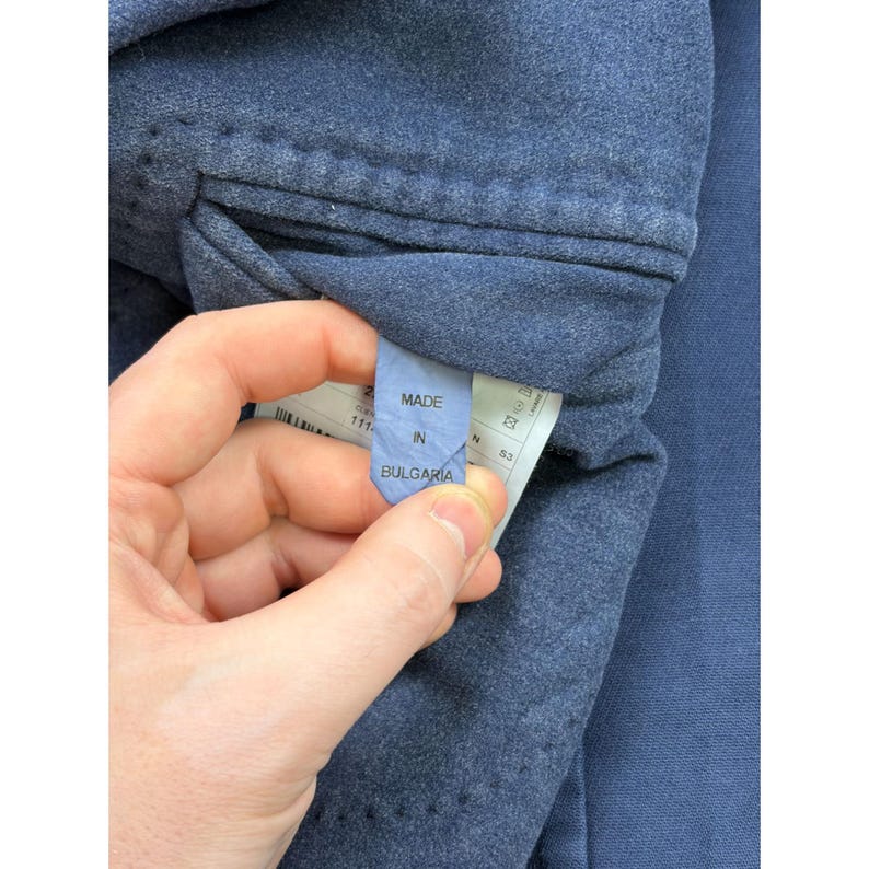 May include: A blue garment featuring an interior fabric label with the text MADE IN BULGARIA printed in black. The label is attached to the inner lining of the blue textile, which displays a textured, slightly mottled appearance throughout.
