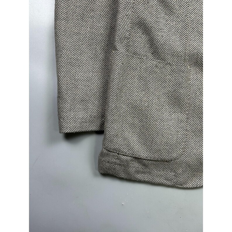 May include: A light gray and white blazer featuring a classic herringbone pattern. The garment includes a visible patch pocket on the front panel and a structured sleeve cuff, showcasing the woven texture of the fabric in a neutral color palette.