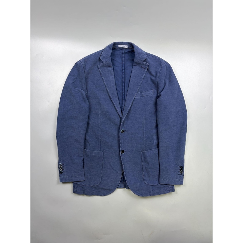 May include: A blue blazer featuring a notched lapel, two-button front closure, and two patch pockets at the waist. The garment includes a single chest pocket, long sleeves with four buttons at each cuff, and visible stitching along the edges.
