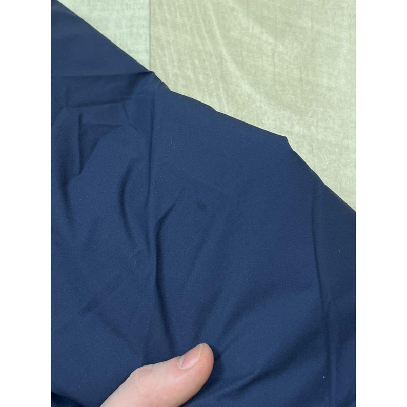 May include: Close-up of a navy blue fabric, possibly a garment. The fabric is smooth and appears to be a solid color. The image is well-lit, showing the texture and folds of the material. A hand is visible in the bottom of the image.
