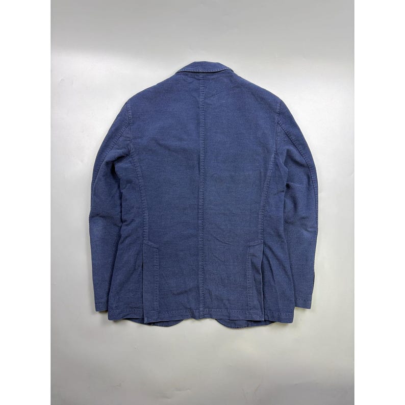 May include: A solid blue blazer shown from the back, featuring a structured collar, long sleeves, and two vertical vents at the lower hem. The garment is crafted from a textured fabric with visible seam detailing along the center back and sides.