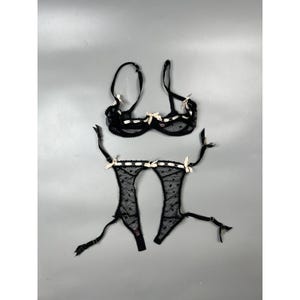 May include: A black sheer bra and matching garter belt set featuring a delicate polka dot pattern. Both pieces are accented with light cream-colored ribbon bows and trim along the edges, including adjustable straps and metal hardware components.