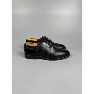 Könnte beinhalten: Ein Paar schwarze Wingtip-Oxford-Schuhe aus Leder mit dekorativen Brogue-Perforationen entlang der Nähte, einer abgerundeten Schuhspitze, schwarzen Schnürsenkeln und einem niedrigen Absatz. In jedem Schuh steckt ein heller Holzschuhspanner.