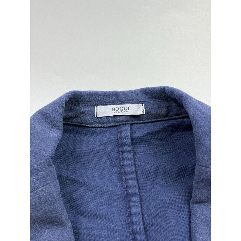 May include: A blue blazer with a textured finish and a notched lapel. The interior collar features a white fabric label with the text Boggi Milano printed in black. The garment has visible stitching along the inner seams and a structured collar.
