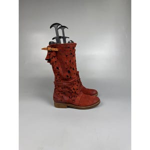 May include: A mid-calf boot in a deep rust-red color, featuring an intricate laser-cut geometric pattern across the shaft. The boot includes a tan leather strap with a metal buckle at the top and a black plastic shoe tree inserted inside.