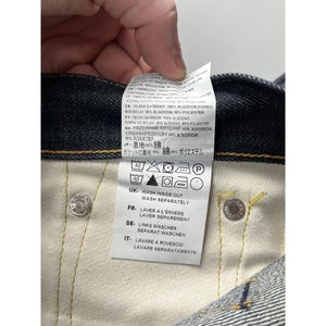 May include: A white fabric care label attached to the interior of dark blue denim jeans. The label features multi-language text detailing material composition, including 100% cotton, and provides laundry instructions with icons and text in English.