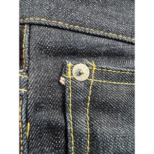 May include: Dark indigo denim fabric featuring a silver-toned metal rivet at the corner of a pocket. The material displays a distinct twill weave pattern and is constructed with thick, contrasting golden-yellow stitching throughout the seams.