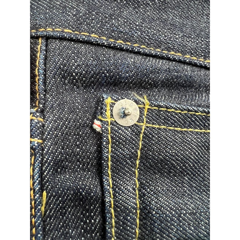 May include: Dark indigo denim fabric featuring a silver-toned metal rivet at the corner of a pocket. The material displays a distinct twill weave pattern and is constructed with thick, contrasting golden-yellow stitching throughout the seams.