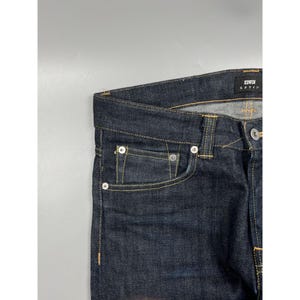 May include: Dark indigo denim jeans featuring a classic five-pocket construction with gold-tone contrast stitching. A black interior label displays the brand name EDWIN in white text. The waistband includes belt loops and a silver-tone metal button.