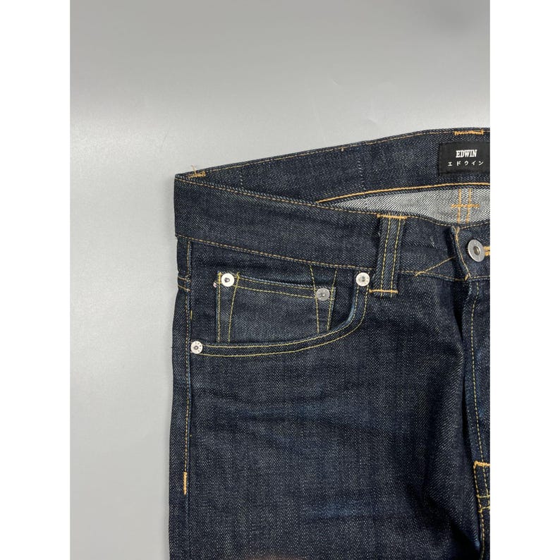 May include: Dark indigo denim jeans featuring a classic five-pocket construction with gold-tone contrast stitching. A black interior label displays the brand name EDWIN in white text. The waistband includes belt loops and a silver-tone metal button.