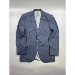 May include: A blue blazer featuring a white pinstripe pattern, notched lapels, and two front buttons. The jacket includes two flap pockets at the waist and a single chest pocket, finished with a light grey interior lining visible at the front opening.