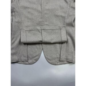 May include: A light gray and white blazer featuring a classic herringbone pattern. The garment displays a structured design with a folded hem detail at the bottom, showcasing the intricate weave of the fabric and the clean, tailored seams.