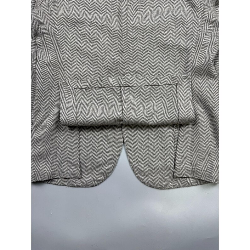 May include: A light gray and white blazer featuring a classic herringbone pattern. The garment displays a structured design with a folded hem detail at the bottom, showcasing the intricate weave of the fabric and the clean, tailored seams.