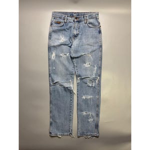 May include: Light blue denim jeans with distressed detailing, featuring multiple rips and tears across the front of the legs. The jeans have a classic five-pocket design and a button and zipper closure. The jeans are a straight-leg style.