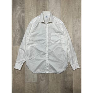 May include: A white long-sleeve shirt with a button-down front and a chest pocket. The shirt has a classic collar and buttoned cuffs. The fabric appears to be lightweight and slightly textured, with visible creases.