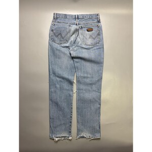 May include: Light blue denim jeans with a distressed look. The jeans feature the Wrangler logo on the back pockets and a brown leather patch. The bottom hems are frayed, and there is a tear on the left leg.