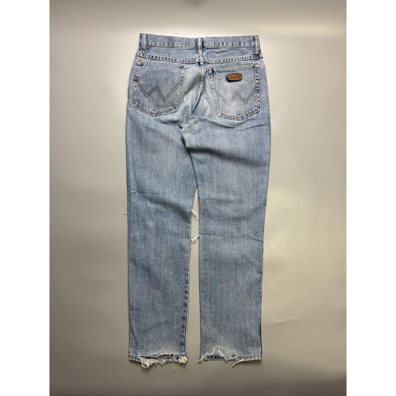 May include: Light blue denim jeans with a distressed look. The jeans feature the Wrangler logo on the back pockets and a brown leather patch. The bottom hems are frayed, and there is a tear on the left leg.