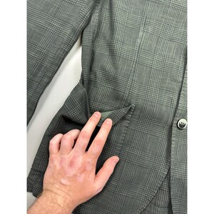 May include: A dark olive green blazer featuring a subtle glen plaid pattern. The garment includes a single visible dark button on the front and a flap pocket detail on the side. The fabric has a textured appearance with a consistent grid design.