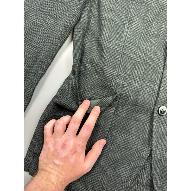 May include: A dark olive green blazer featuring a subtle glen plaid pattern. The garment includes a single visible dark button on the front and a flap pocket detail on the side. The fabric has a textured appearance with a consistent grid design.