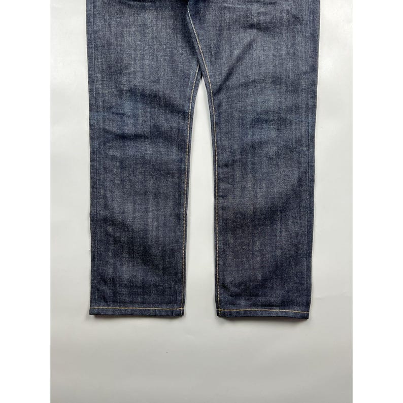 May include: A pair of dark indigo blue denim jeans featuring a straight leg cut, visible textured fabric weave, and contrasting gold-toned stitching along the side seams and the bottom hemline of both pant legs, showcasing a classic construction.