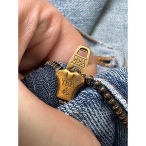 May include: Close-up of a brass-colored YKK zipper on blue denim fabric. The zipper pull is open, revealing the teeth and the YKK brand name and the number 45.