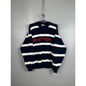 May include: A long-sleeve crewneck sweatshirt featuring a horizontal striped pattern in navy blue and white. The word BENETTON is embroidered in red capital letters across the chest. The garment has a ribbed collar, cuffs, and hemline.