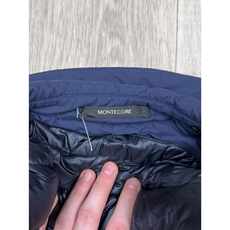 May include: Close-up of a navy blue jacket with a black interior. The jacket's label reads "MONTECORE" in white text on a black background. The jacket is partially open, revealing the quilted lining.