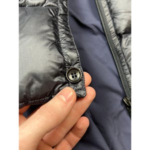 May include: Close-up of a dark navy blue quilted jacket. The jacket features a button closure and a zipper. The interior lining is a darker shade of blue. The jacket appears to be made of a smooth, shiny material.