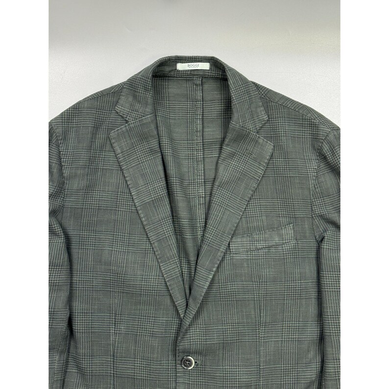 May include: A dark gray blazer featuring a subtle black plaid pattern, notched lapels, a single-button front closure, and a chest pocket. The interior collar label displays the text Boggi Milano in black lettering on a white background.