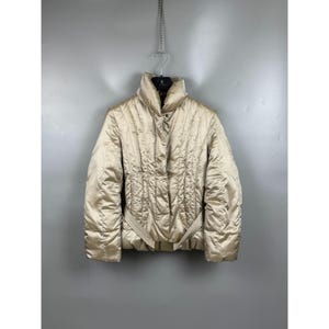 Könnte beinhalten: Eine champagnerfarbene Steppjacke mit hohem Kragen, langen Ärmeln und einem Frontreißverschluss, der von einer Druckknopfleiste verdeckt wird. Die Jacke verfügt über vertikale Steppnähte, eine taillierte Passform und einen passenden Gürtel.