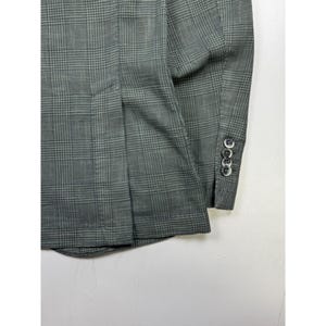 May include: A grey blazer featuring a dark grey glen plaid pattern. The garment includes a long sleeve with four small, round, dark-colored buttons aligned vertically along the cuff and a vertical seam detail on the side of the torso.