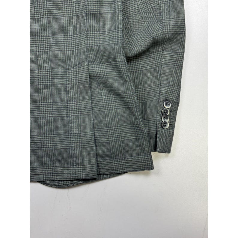 May include: A grey blazer featuring a dark grey glen plaid pattern. The garment includes a long sleeve with four small, round, dark-colored buttons aligned vertically along the cuff and a vertical seam detail on the side of the torso.