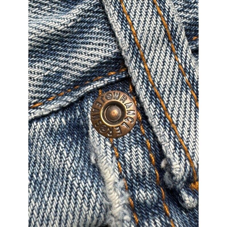 May include: Close-up of blue denim fabric with a bronze-colored button. The button is embossed with the text "ROW RANGE JEANS". The denim has visible stitching in a contrasting gold color.
