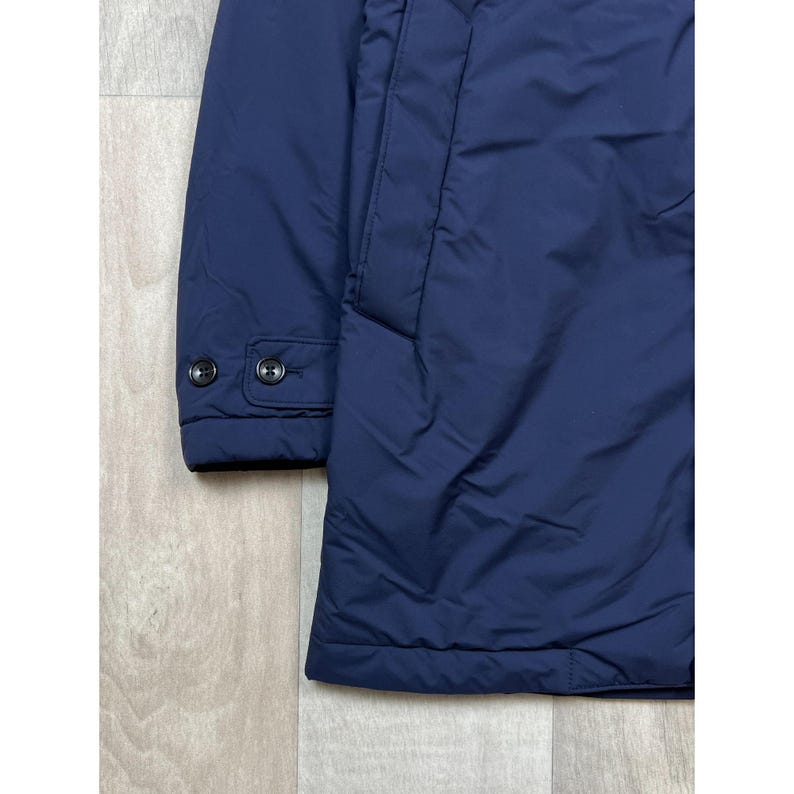May include: A navy blue jacket with a buttoned cuff and two black buttons. The jacket has a quilted appearance and a front pocket. The fabric has a slight sheen, suggesting a water-resistant material.