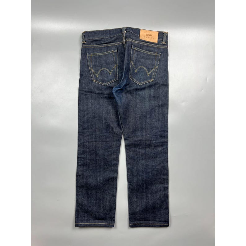 May include: Dark indigo denim jeans featuring a tan leather patch on the back waistband with the text EDWIN printed in black. The jeans have two back pockets with yellow contrast stitching in a curved arc design and belt loops at the waist.
