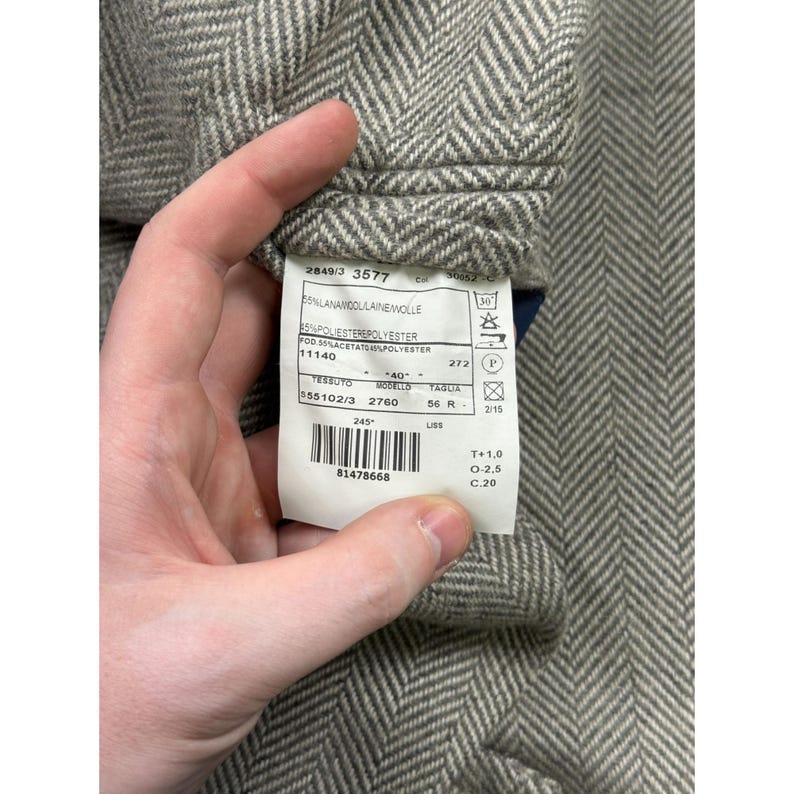 May include: A grey and white herringbone patterned fabric with a white rectangular garment care label attached. The label displays text including 55% Lana Wool, 45% Polyester, 55% Acetate, model number 2760, size 56 R, and a barcode.