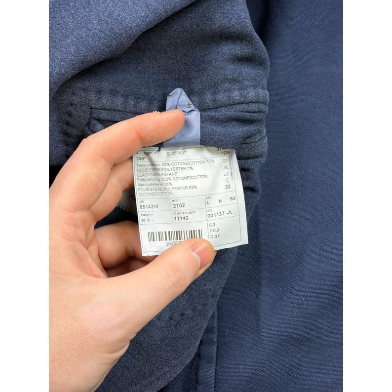 May include: A dark blue garment featuring an interior white fabric care label. The label displays text including material composition of 82% cotton, 17% polyester, and 1% elastane, along with a barcode and various product identification codes.