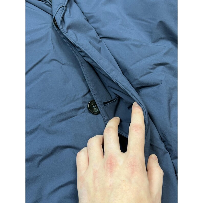 May include: Close-up of a navy blue jacket with a buttoned pocket. The jacket has a smooth, slightly shiny texture. A hand is reaching into the pocket, showing the inside lining. The button is black and round.