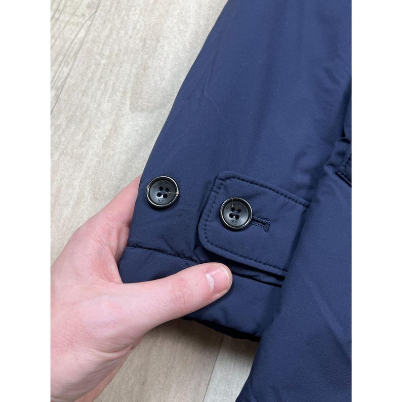 May include: Close-up of a navy blue jacket sleeve with two black buttons. The sleeve has a buttoned cuff detail. The jacket appears to be made of a smooth, possibly water-resistant material.