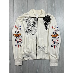 May include: Cream-colored zip-up jacket with black and red tattoo-style graphics. Features an eagle, skull, and dagger designs, along with the text "New York City" and "Slowly". The jacket has a collared neckline and two front pockets.