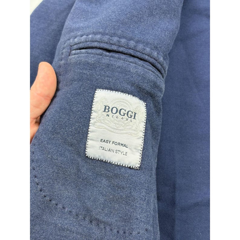 May include: A navy blue blazer featuring a textured fabric and a rectangular white interior label. The label displays the text BOGGI MILANO, EASY FORMAL, and ITALIAN STYLE in dark blue lettering, positioned near a welt pocket on the inner lining.