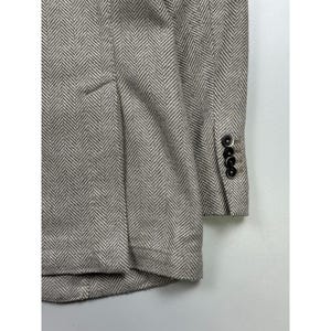May include: A beige and white blazer featuring a prominent herringbone pattern throughout the fabric. The garment includes a structured sleeve cuff detailed with four small, dark circular buttons arranged in a vertical row along the side.