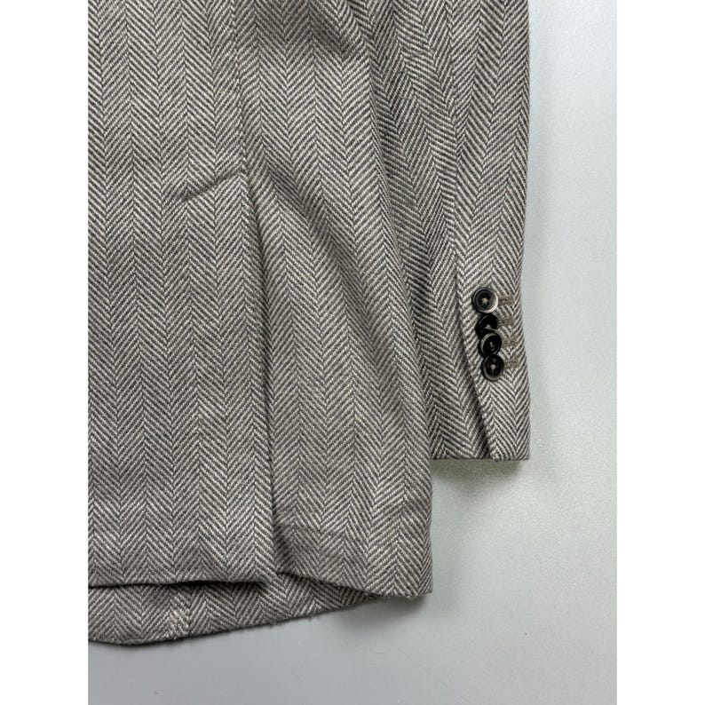May include: A beige and white blazer featuring a prominent herringbone pattern throughout the fabric. The garment includes a structured sleeve cuff detailed with four small, dark circular buttons arranged in a vertical row along the side.