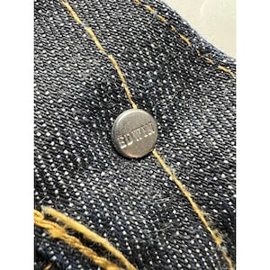 May include: A dark blue denim fabric featuring a metallic silver button embossed with the text EDWIN. The denim is constructed with thick, golden-yellow contrast stitching that runs vertically across the textured, woven surface of the material.