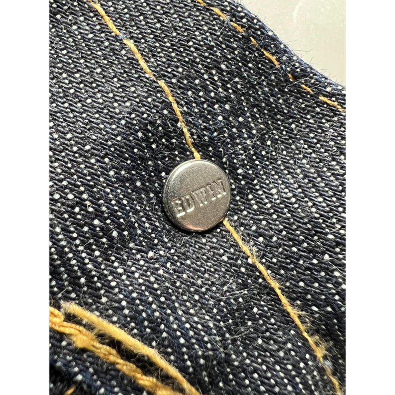May include: A dark blue denim fabric featuring a metallic silver button embossed with the text EDWIN. The denim is constructed with thick, golden-yellow contrast stitching that runs vertically across the textured, woven surface of the material.
