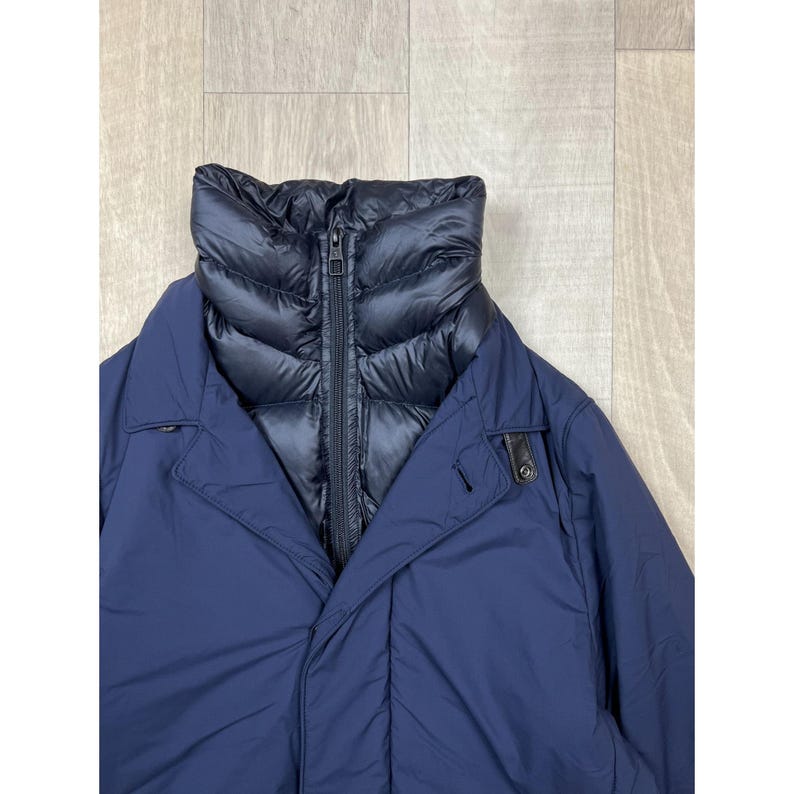 May include: A navy blue winter coat with a black, quilted inner layer and a high collar. The coat has a zipper closure and a button detail on the front. The outer layer is smooth, while the inner layer has a puffy texture.