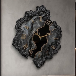 Puede incluir: Un gran reloj de pared decorativo con un diseño abstracto único. El reloj presenta un marco texturizado negro y gris con números romanos y manecillas doradas. Cuelga sobre un sofá beige y una mesa de centro negra en un salón moderno.