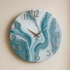 Può includere: Un orologio da parete rotondo con quadrante bianco e design marmorizzato blu e bianco. Lancette e numeri argentati indicano l'ora. L'orologio è montato su una parete beige sopra un divano beige con cuscini blu e beige.
