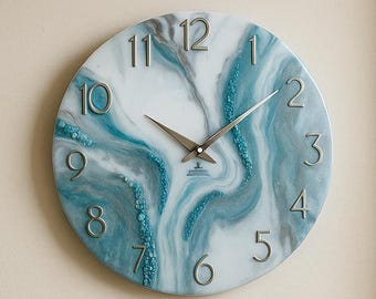 May include: A round wall clock with a white face and blue and white marbled design. Silver-colored hands and numerals mark the time. The clock is mounted on a beige wall above a beige sofa with blue and beige pillows. A small potted plant sits nearby.